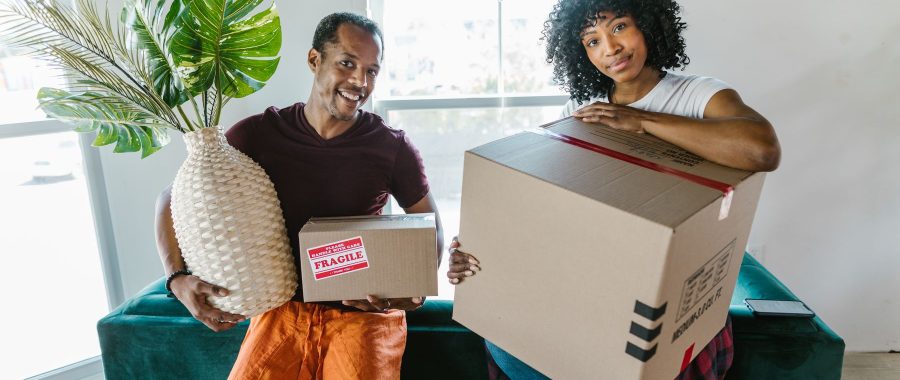 Un couple souriant emménage dans un logement lumineux, un carton et une plante dans les bras