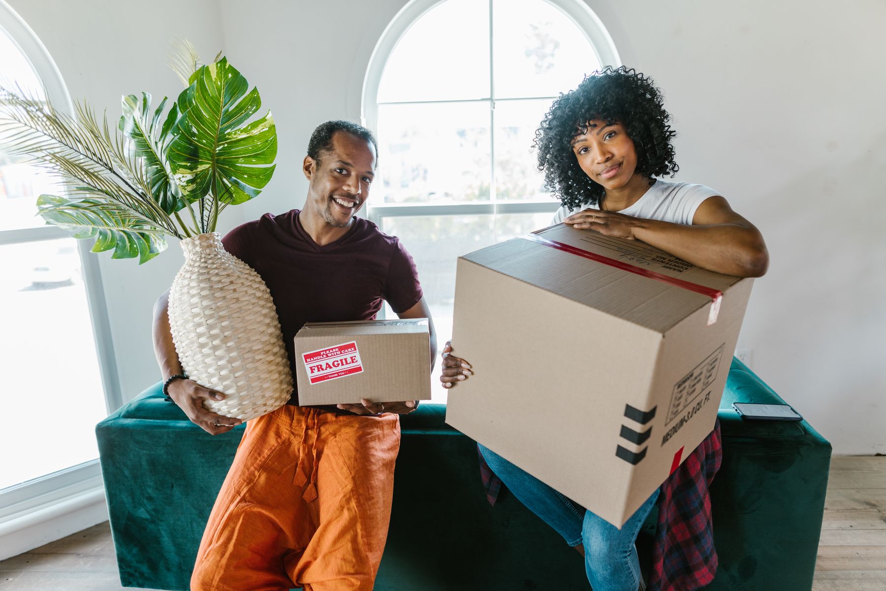 Un couple souriant emménage dans un logement lumineux, un carton et une plante dans les bras
